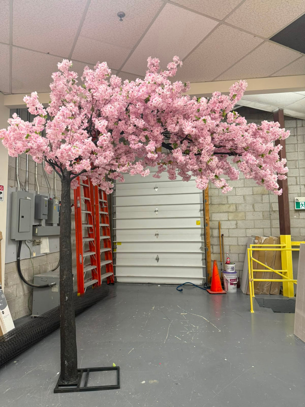 An artificial white pink cherry blossom  arch tree with dense pink flowers and a dark trunk, displayed in a warehouse or garage setting with a gray floor