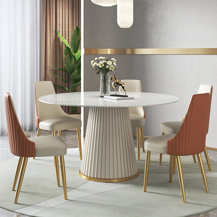 Elegant dining room with a modern round table featuring a fluted base and marble top, complemented by five stylish chairs with beige and orange upholstery and gold legs, set against a chic gray and gold-accented wall with a touch of greenery.
