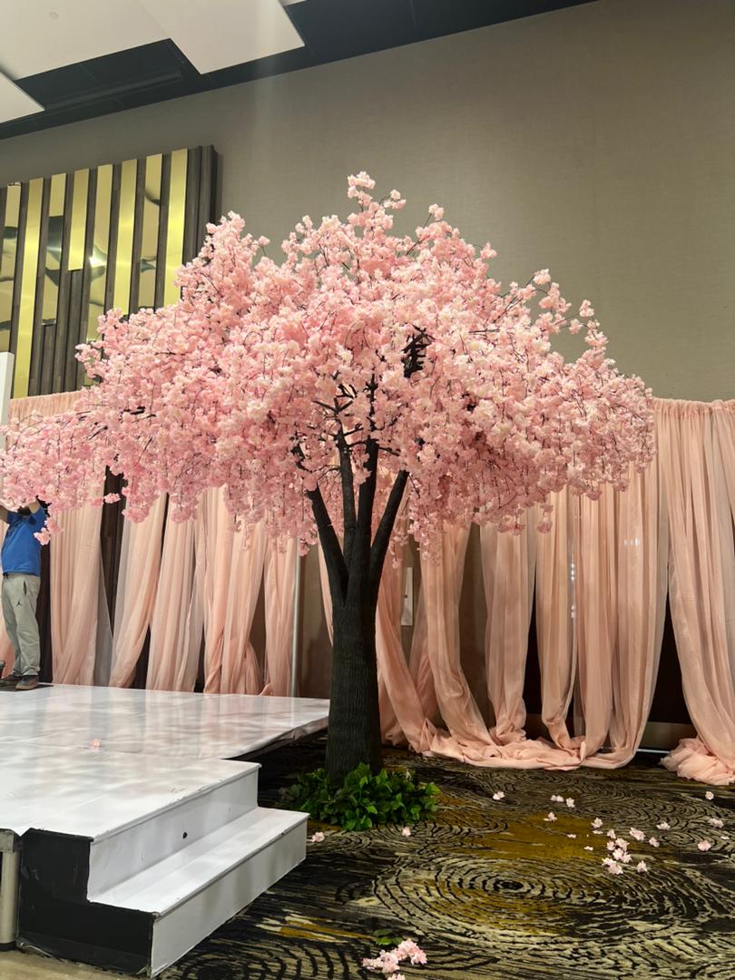 A 15ft pink weeping artificial pink cherry blossom tree displayed indoors in front of draped blush pink fabric, with a white platform and patterned carpet visible.