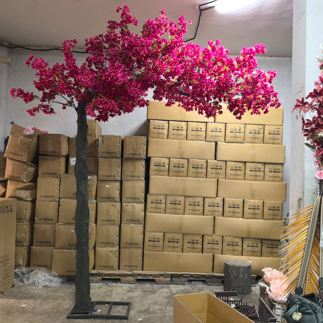 Artificial bougainvillea tree with vibrant pink flowers displayed in a storage room. Background features stacks of cardboard boxes and decorative items, highlighting the tree's potential for event décor setups.
