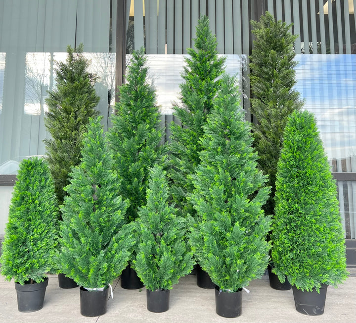 Collection of artificial evergreen trees in varying sizes, all potted in black containers, arranged outdoors against a window, ideal for creating a lush, natural ambiance in any setting.