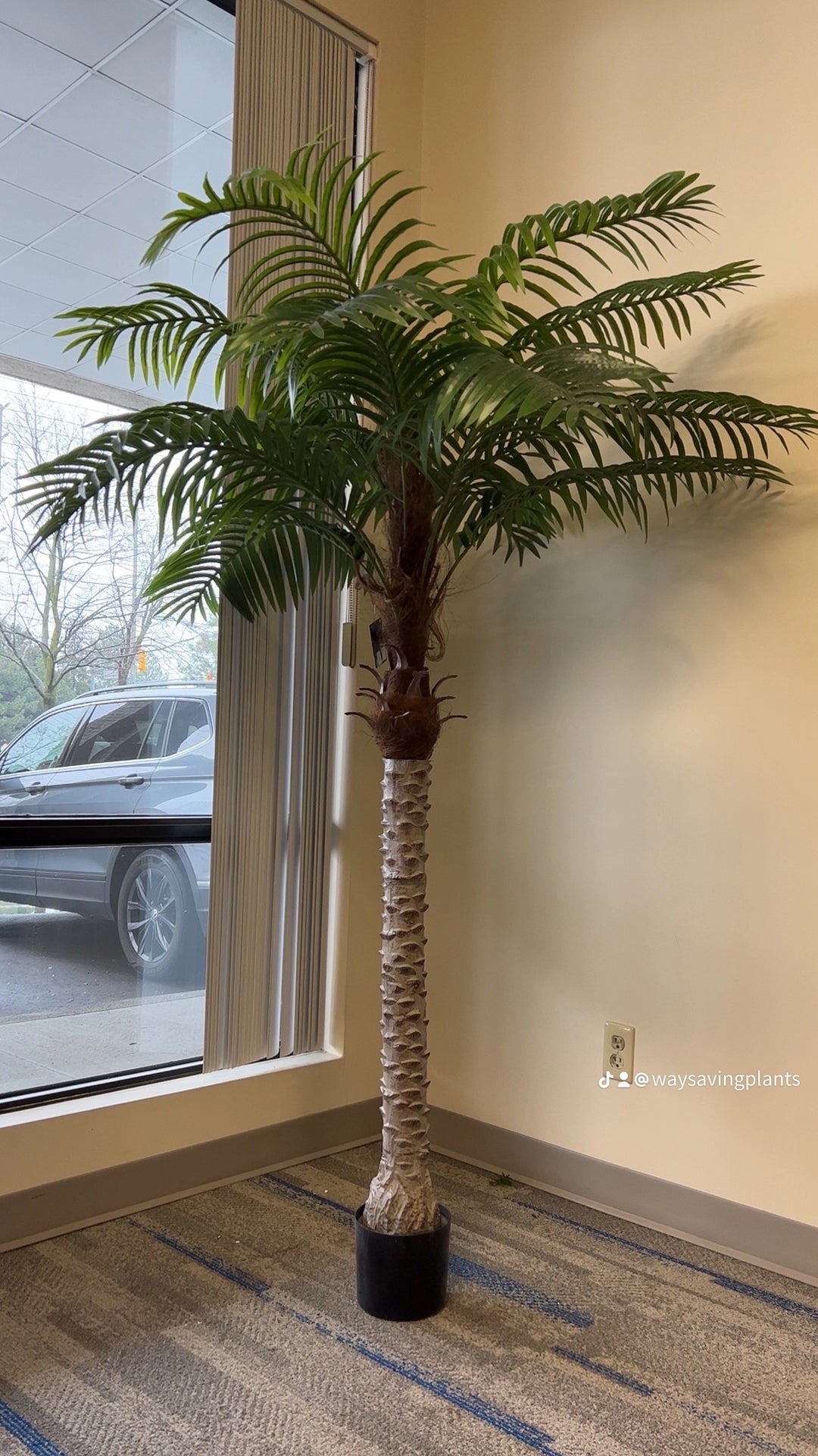 A large artificial palm tree in a stylish office corner by the window, adding a natural touch to a professional setting.