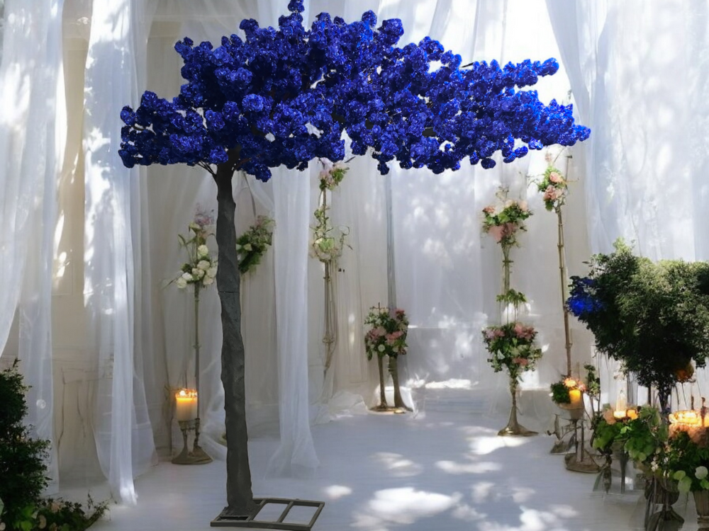 A vibrant blue artificial tree is set against an ethereal backdrop of flowing white drapery in an elegant venue. The space is adorned with floral arrangements in soft pastel tones, complemented by golden stands and glowing candles, creating a romantic and serene ambiance.