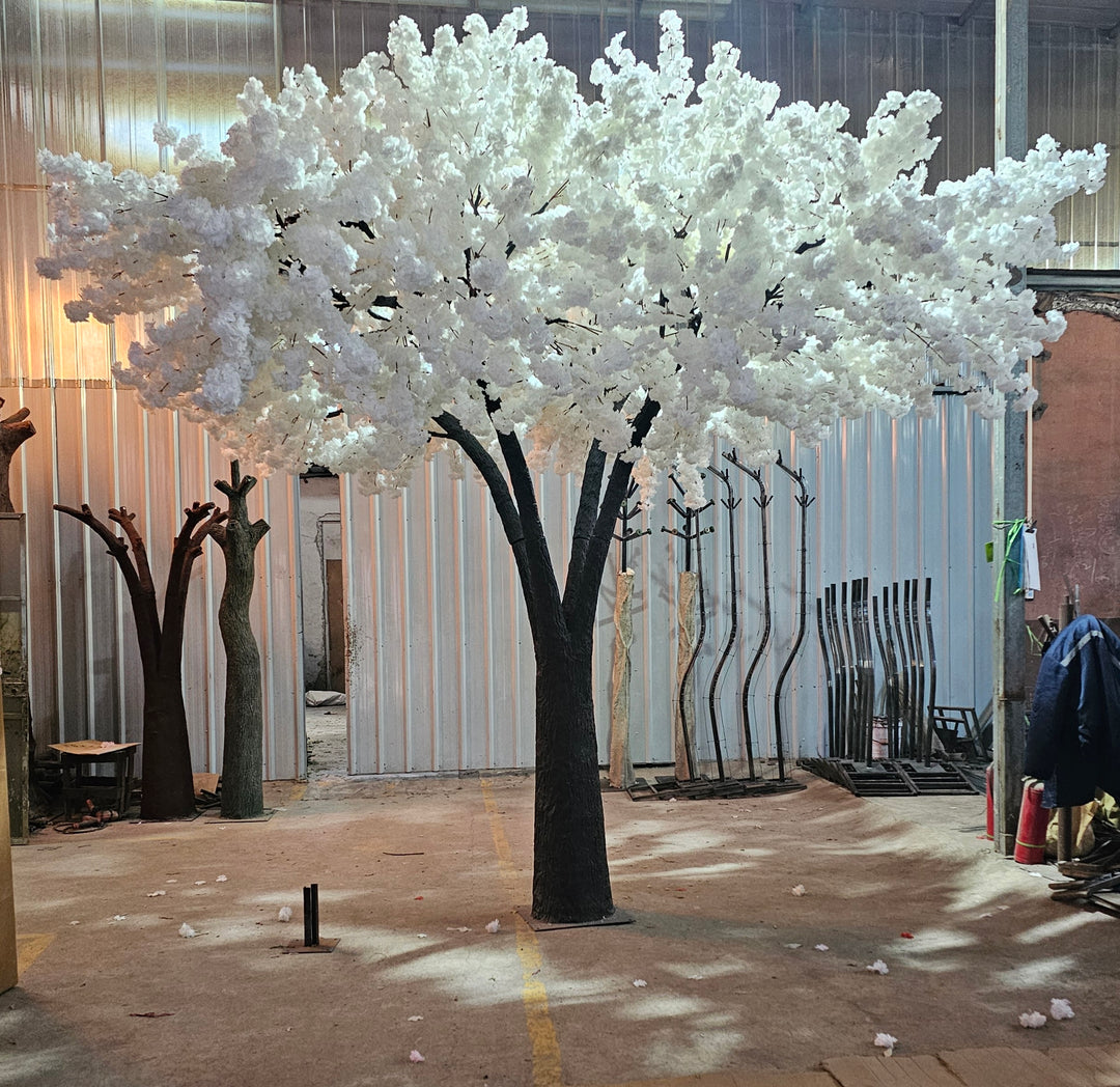 Artificial tree with lush white blossoms and a realistic trunk, displayed in a workshop setting, ready for rental as stunning decor for weddings and events. Are these trees easy to assemble and transport for venue setup?