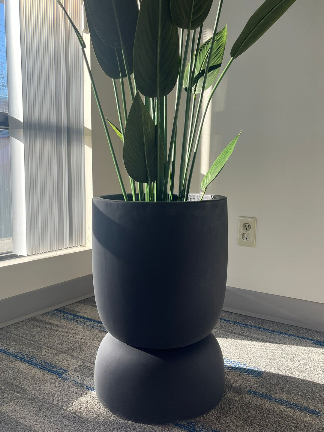 Close-up of a black stackable planter with a smooth matte finish, holding a tall artificial plant with large green leaves, placed indoors near a window with natural light. Does this planter have drainage holes?