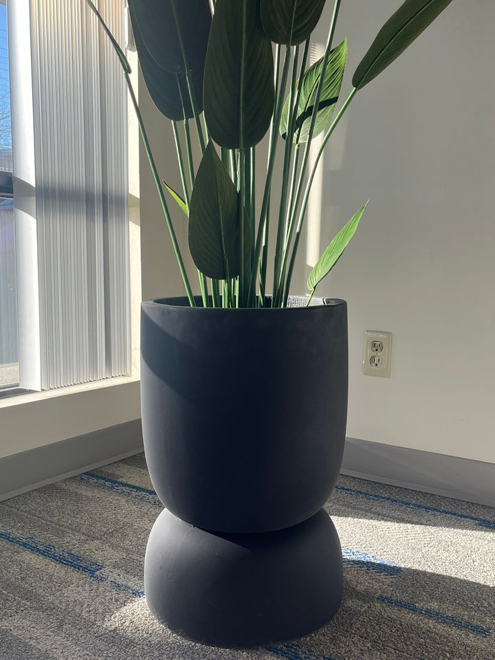 Close-up of a black stackable planter with a smooth matte finish, holding a tall artificial plant with large green leaves, placed indoors near a window with natural light. Does this planter have drainage holes?