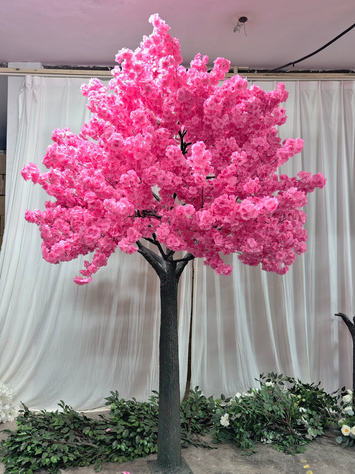 A vibrant artificial pink cherry blossom tree displayed indoors against a white curtain backdrop. The tree has a sturdy dark trunk and is surrounded by scattered green leaves and floral branches on the floor, showcasing its realistic and decorative design.