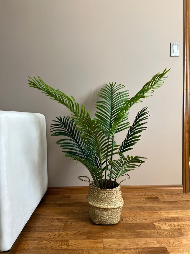 Artificial palm plant in a woven basket, placed on a wooden floor beside a white sofa, enhancing the natural and cozy interior space.