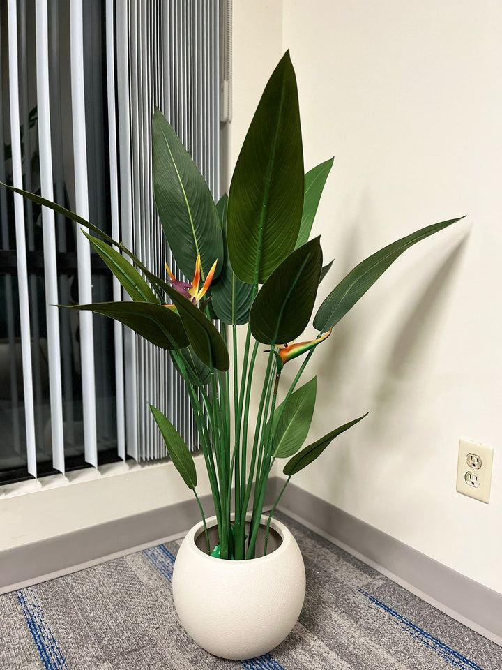 Ivory round planter with a smooth matte finish, holding a vibrant artificial bird of paradise plant with broad green leaves and colorful flowers, placed indoors near a window. Does this planter come with a protective inner lining?