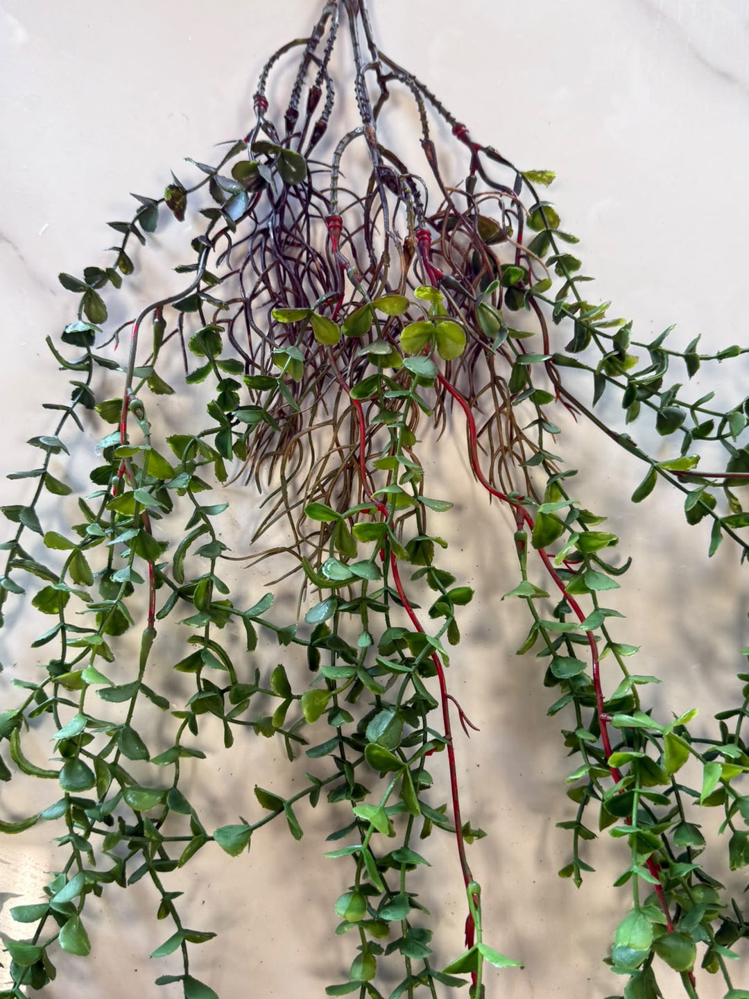 A hanging artificial trailing plant with small green leaves and reddish stems, attached to a bundle of dark brown faux roots, shown against a light neutral background.