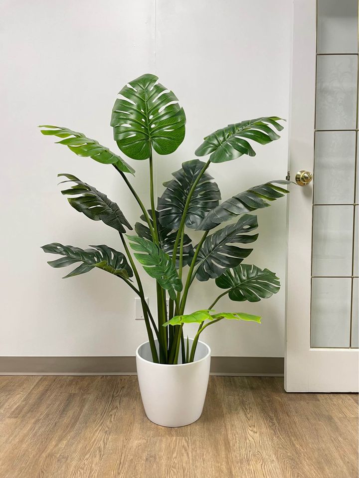 Large artificial monstera plant with vibrant green, split leaves, displayed in a sleek white pot on a wooden floor, enhancing the indoor decor with a tropical touch