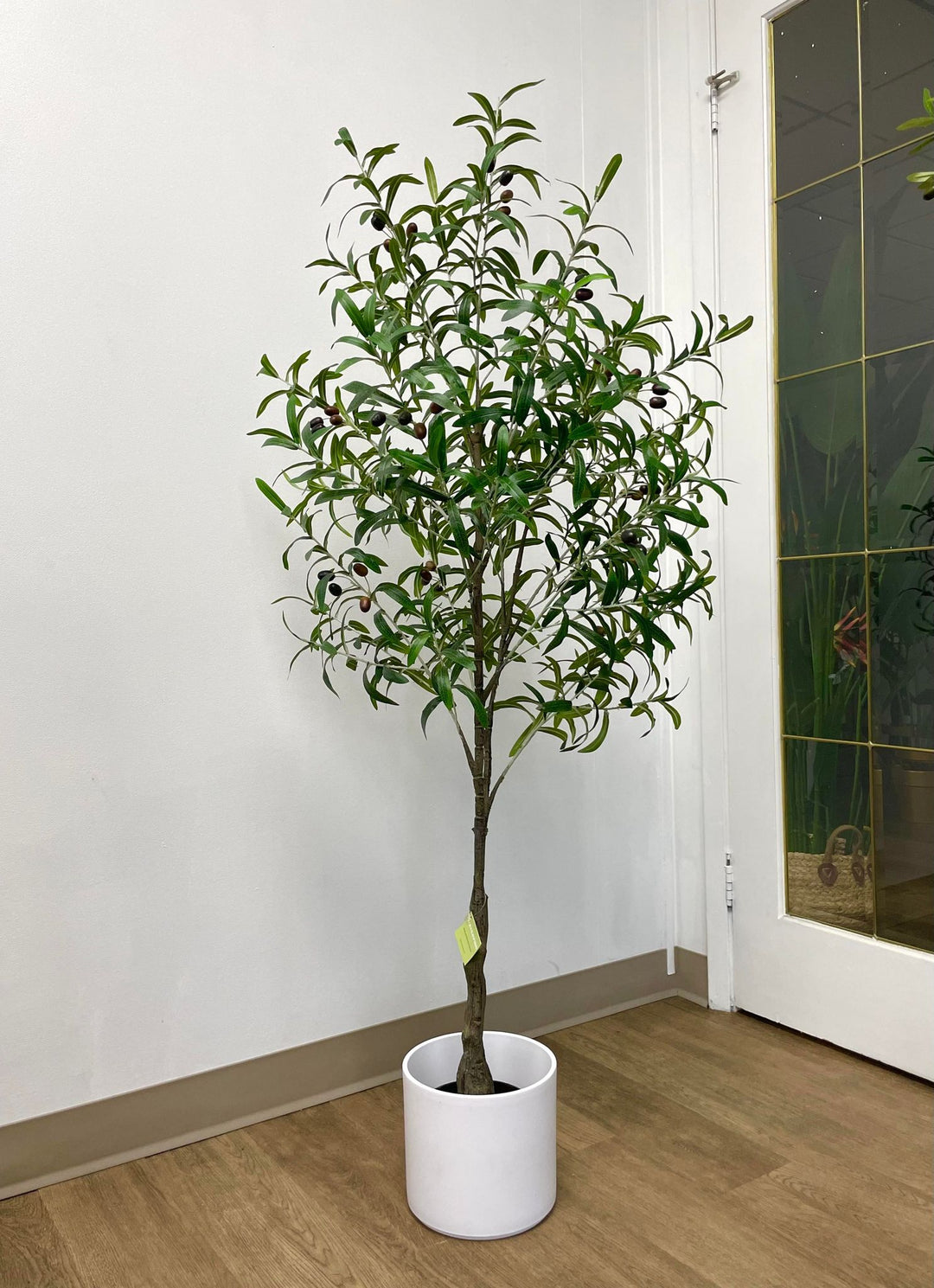Artificial olive tree placed in a textured, light brown pot with a leaf pattern. The tree is positioned near a window with vertical blinds, filling the corner of a room with a neutral wall color and carpeted floor.