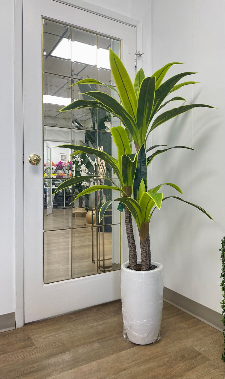 Elegant tall artificial plant with vibrant green leaves in a sleek white pot, placed in a modern indoor setting with reflective glass panels for a chic home or office decor.
