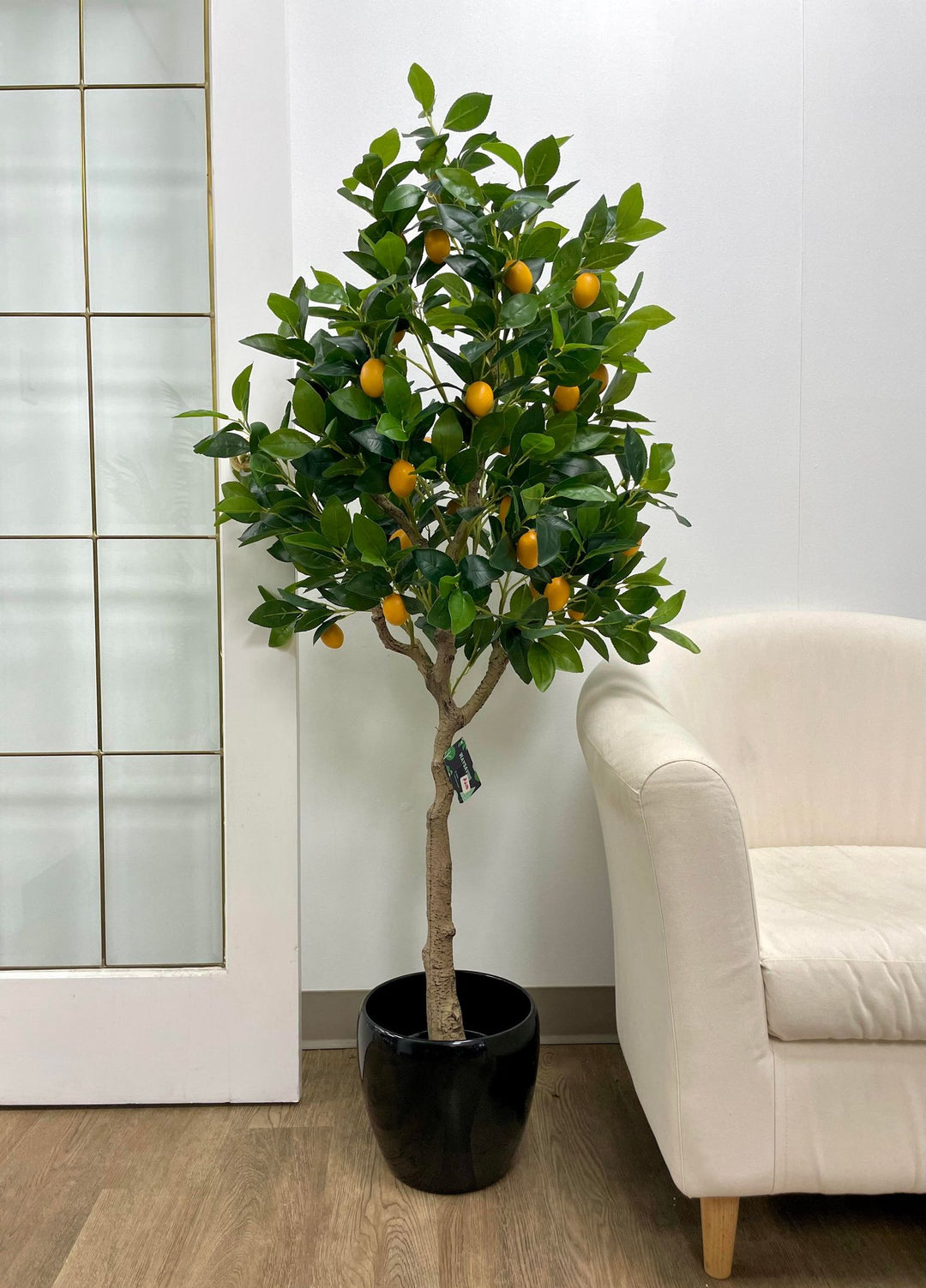 Artificial lemon tree with lush green leaves and bright yellow lemons in a tall black planter, placed near a glass-paneled door in a modern indoor setting
