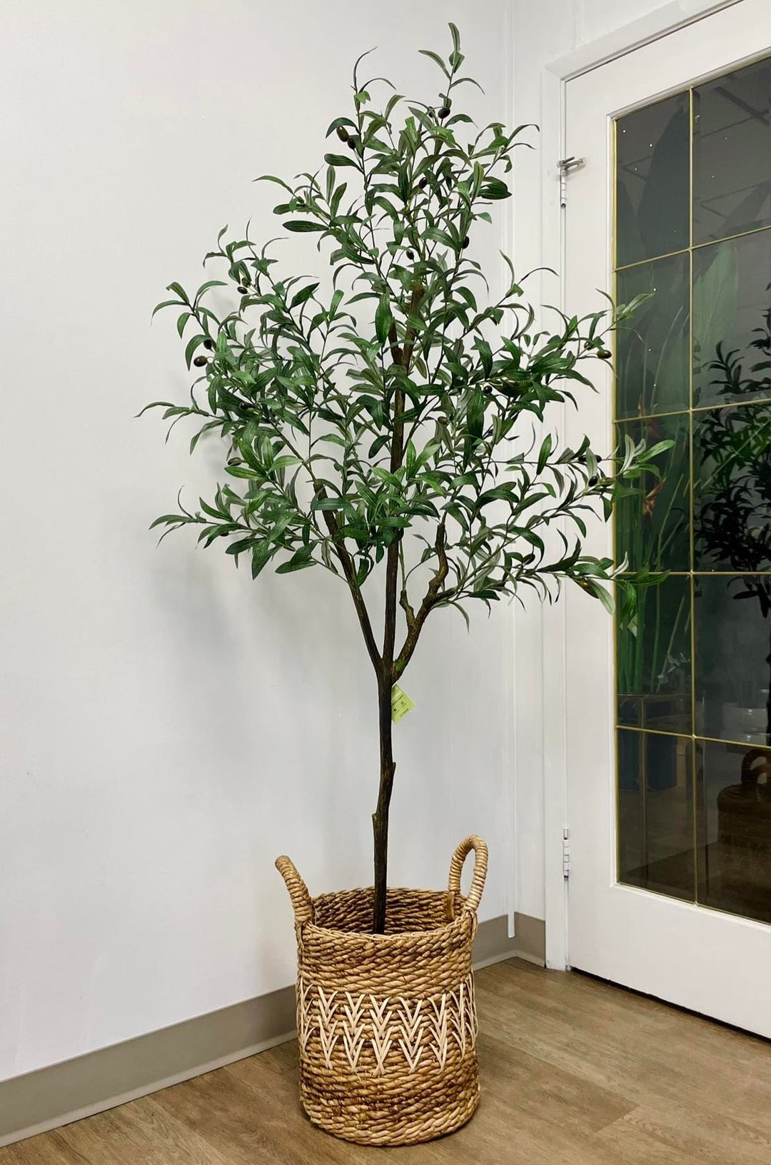 Artificial olive tree with slender green leaves and small black olives in a textured white pot placed near a glass-paneled door in a bright indoor space with wooden flooring