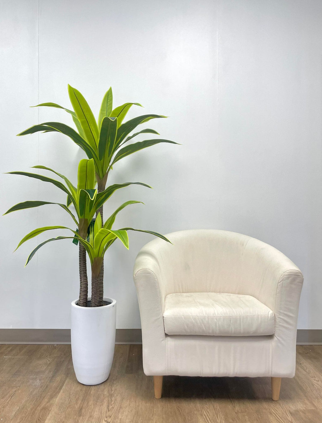 Artificial tall green plant with broad, vibrant leaves in a sleek white pot, paired with a modern cream armchair, perfect for contemporary home or office decor