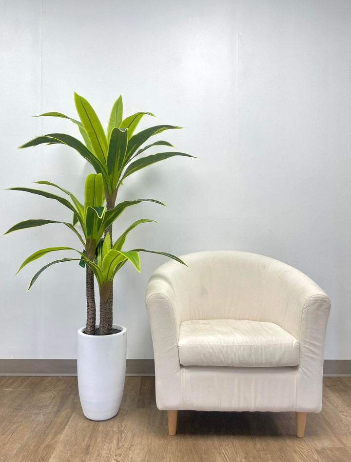 Artificial tall green plant with broad, vibrant leaves in a sleek white pot, paired with a modern cream armchair, perfect for contemporary home or office decor