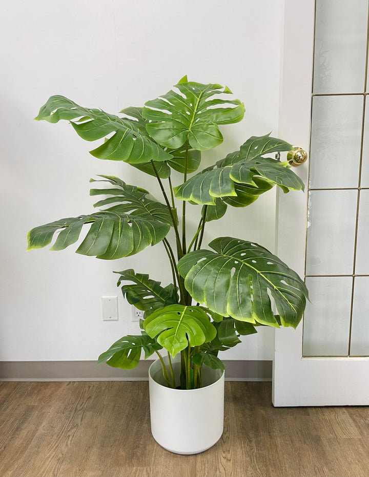 Artificial monstera plant in a white ceramic pot with a wooden stand, positioned next to a cream-colored armchair. The plant features large, realistic green leaves with natural splits, adding a tropical vibe to the indoor space. The setup is placed on a carpeted floor with subtle blue accents
