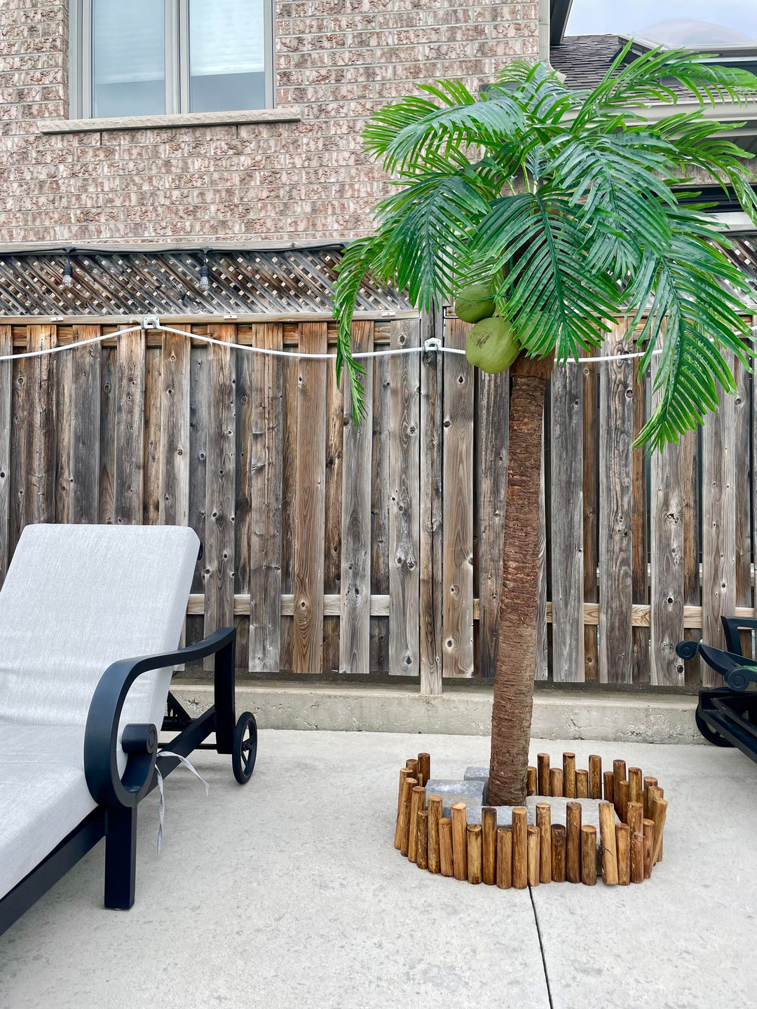 The image shows a patio with a single artificial palm tree, coconuts, a small wooden fence at its base, and a grey lounge chair on a concrete surface. A wooden fence and brick house are in the background.