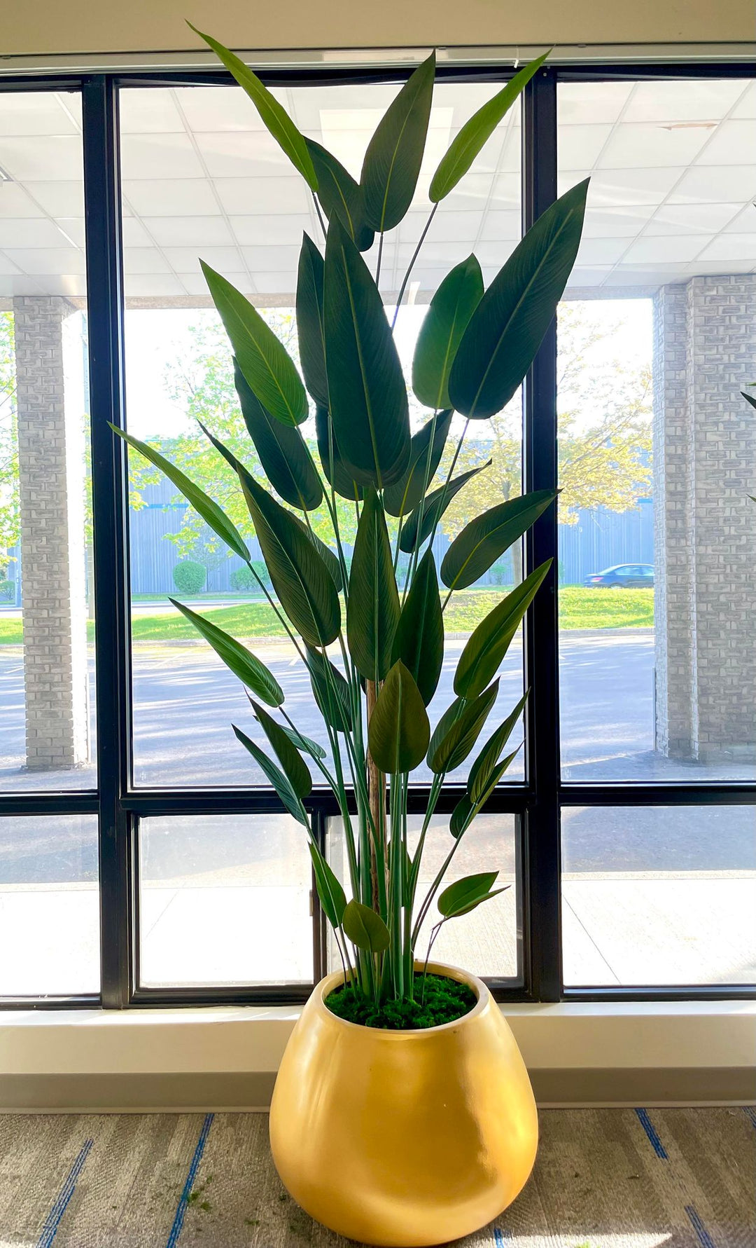 A large artificial bird of paradise plant with tall, green leaves arranged in a golden round pot. The plant is placed in front of a large window, with sunlight filtering through, creating a serene indoor environment