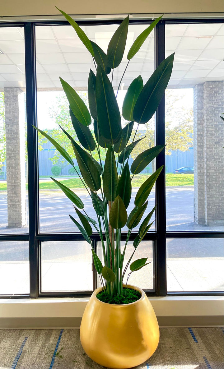 A large artificial bird of paradise plant with tall, green leaves arranged in a golden round pot. The plant is placed in front of a large window, with sunlight filtering through, creating a serene indoor environment