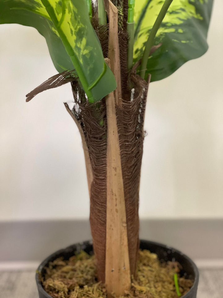 A close-up of the base of a plant showing its textured trunk and the base of the leaves, giving a detailed look at the realistic design and intricate details of the artificial plant. The plant is set in a pot with moss covering the soil, adding a natural touch to the overall appearance.