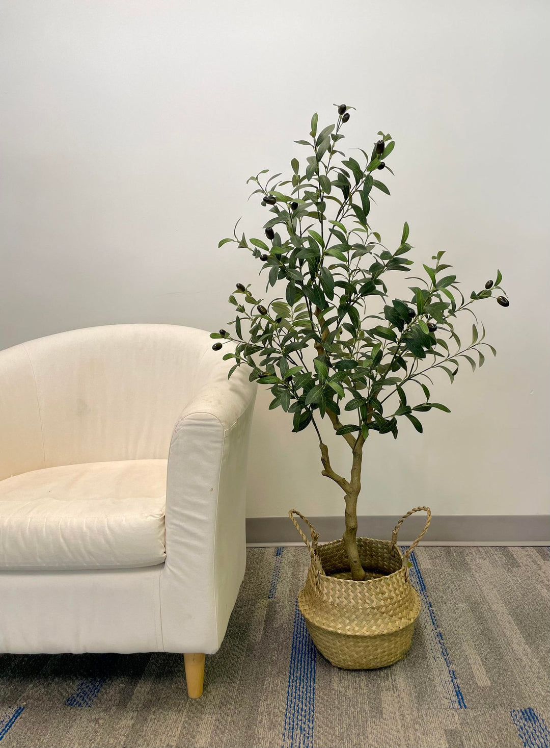 Artificial olive tree in a woven basket, placed next to a white armchair against a light-colored wall on a carpeted floor