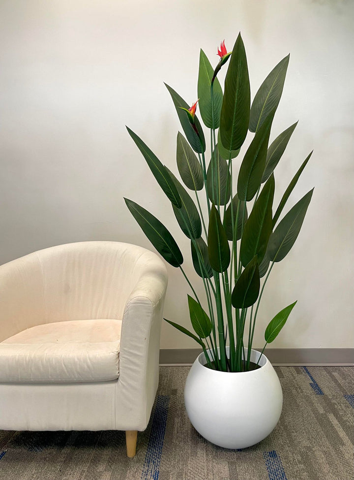 Artificial bird of paradise plant with vibrant green leaves and bright orange flowers in a modern white pot, placed next to a beige armchair, creating a serene and stylish corner in an indoor setting