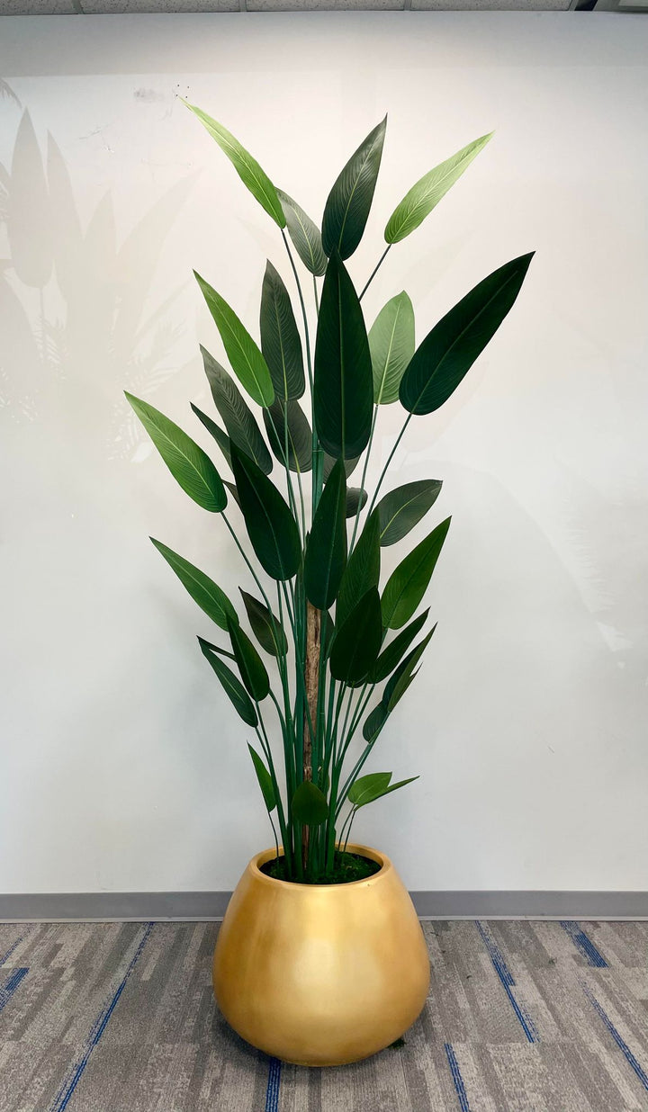 A large artificial bird of paradise plant with tall, green leaves arranged in a golden round pot. The plant is placed in front of a large window, with sunlight filtering through, creating a serene indoor environment