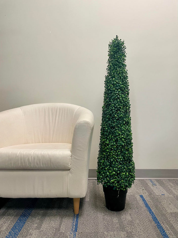A tall, artificial topiary in the shape of a cone stands in a black pot on a gray carpet. The topiary is positioned next to a white upholstered chair, providing a contrast between the soft texture of the chair and the dense, leafy surface of the topiary