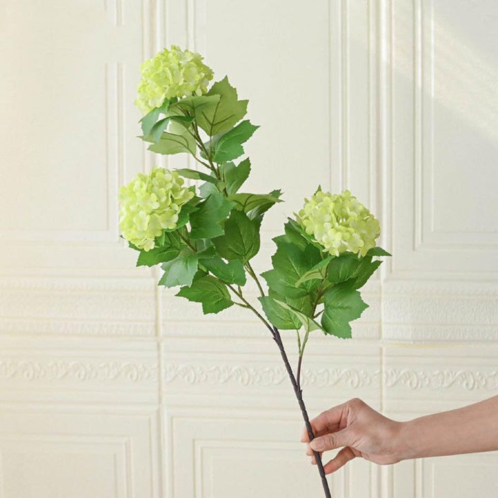A hand holding an artificial hydrangea stem with three green flower clusters. Commonly searched question: How do you decorate with artificial stems to make them look real?