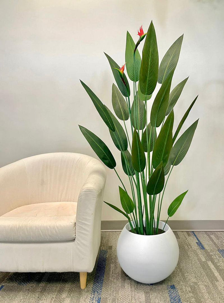 Artificial bird of paradise plant with vibrant green leaves and bright orange flowers in a modern white pot, placed next to a beige armchair, creating a serene and stylish corner in an indoor setting.