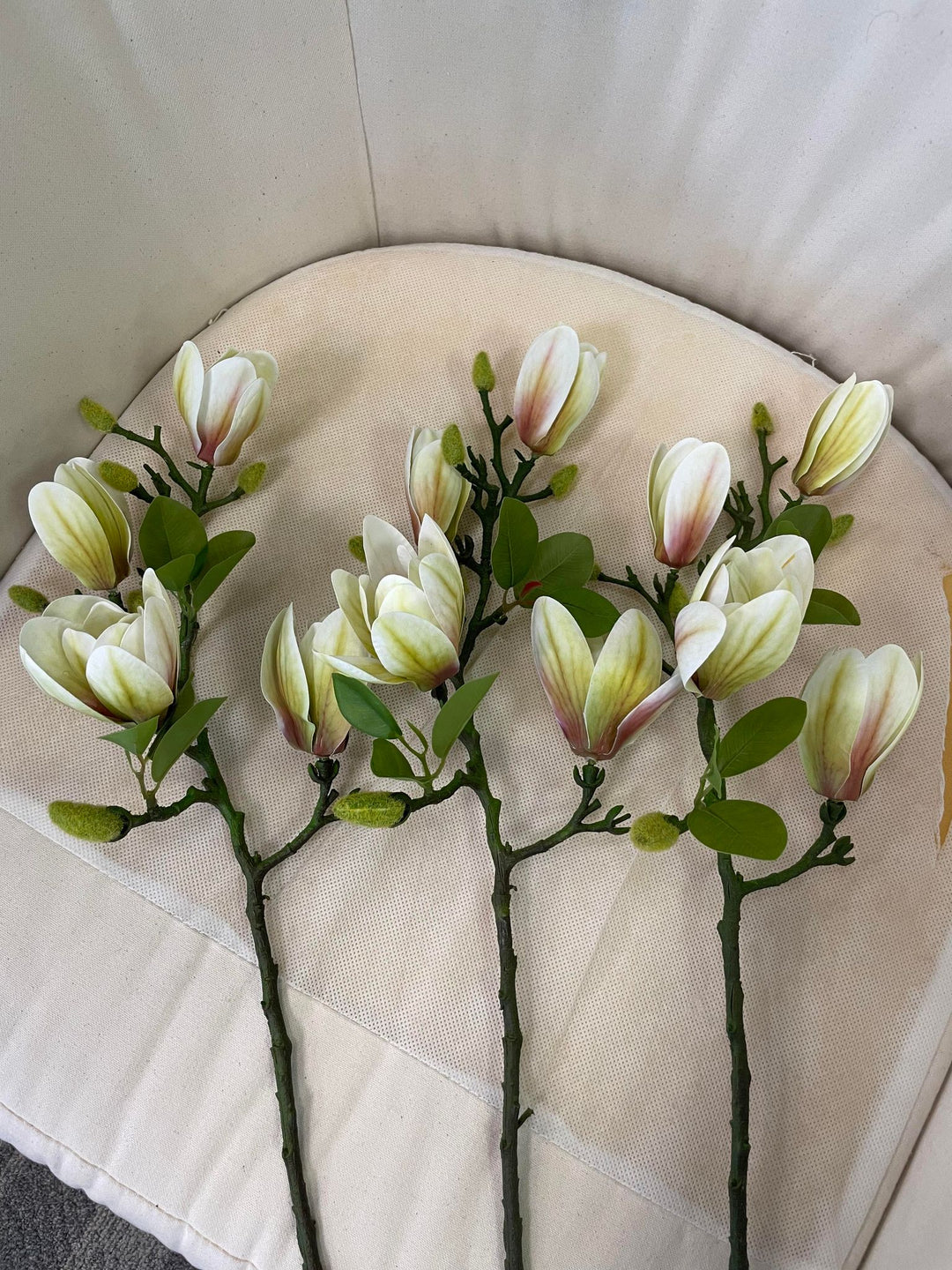 A set of artificial magnolia stems with delicate cream and blush pink flowers laid out on a soft beige chair. Frequently asked question: Are artificial flower stems easy to arrange for DIY home decor projects?"
