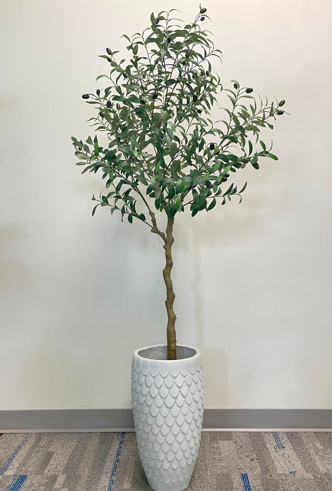 Artificial olive tree placed in a textured, light brown pot with a leaf pattern. The tree is positioned near a window with vertical blinds, filling the corner of a room with a neutral wall color and carpeted floor