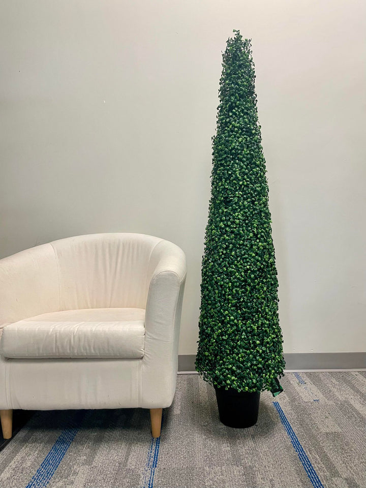 A tall, artificial topiary in the shape of a cone stands in a black pot. The topiary is lush with dense, small green leaves and is placed outside against a textured concrete wall. In the background, there is a view of a building with large windows.