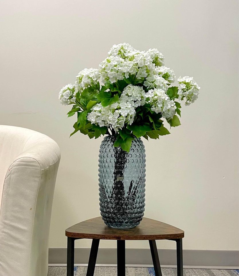 A large arrangement of artificial white hydrangeas in a textured glass vase placed on a small wooden coffee table beside a cream-colored chair. This elegant floral arrangement adds a touch of sophistication to living spaces without the need for maintenance. What are the best artificial flower arrangements for modern home decor?