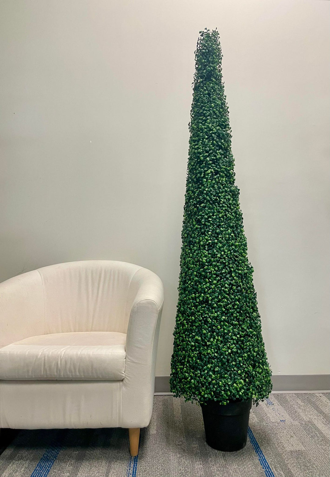A tall, artificial topiary in the shape of a cone stands in a black pot on a gray carpet. The topiary is positioned next to a white upholstered chair, providing a contrast between the soft texture of the chair and the dense, leafy surface of the topiary.