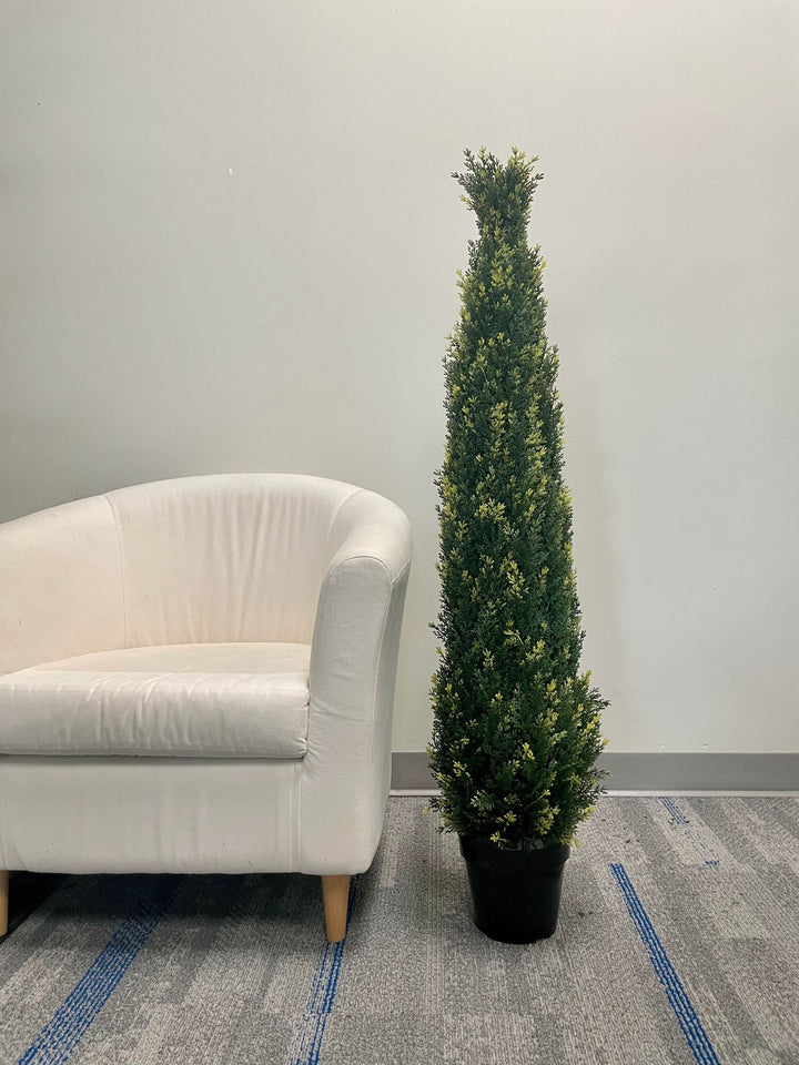 A tall, artificial topiary in the shape of a cone stands in a black pot. The topiary is lush with dense, small green leaves and is placed outside against a textured concrete wall. In the background, there is a view of a building with large windows.