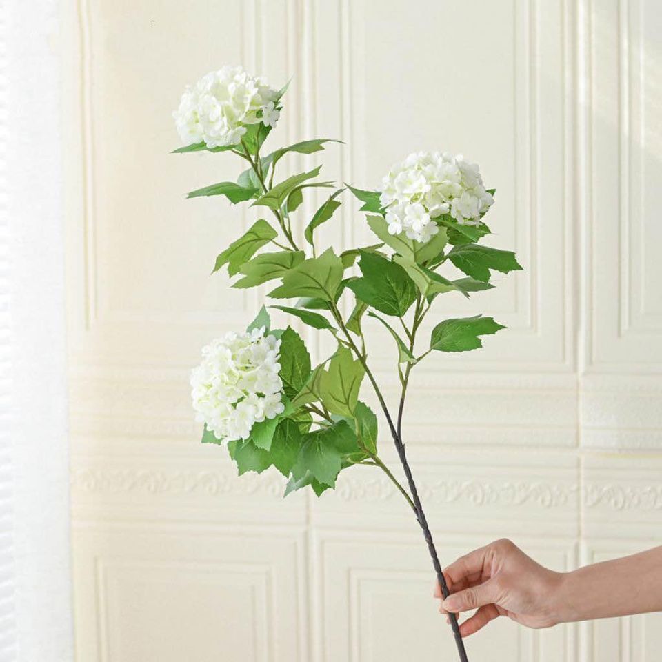 Artificial white hydrangea stem with lush green leaves, held by a hand against a soft beige background. Ideal for home decor arrangements or DIY floral projects, these stems offer a natural look without the maintenance. What are the best artificial plants for low-maintenance decor?