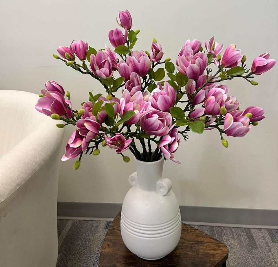 A vibrant arrangement of artificial magnolia flowers in full bloom placed in a white ceramic vase on a small wooden table. The flowers display rich pink and purple tones with green leaves, positioned next to a soft cream armchair. How to decorate your living room with artificial flowers for a fresh look?