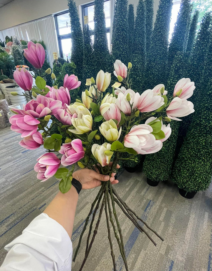 A person holding a large bouquet of artificial magnolia flowers in shades of pink, cream, and white. The vibrant arrangement is set against a backdrop of lush artificial greenery and topiary displays, showcasing how to use faux florals for home or office decor.