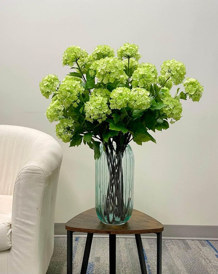 A bouquet of artificial green hydrangeas in a tall transparent vase placed on a triangular wooden coffee table next to a white armchair. What are the best coffee table flower arrangements for a modern living room.