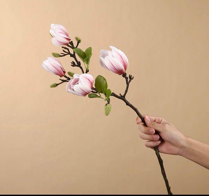 A hand holding a single artificial magnolia branch with pink and white blossoms and fresh green leaves, showcasing a delicate floral arrangement against a soft beige background. Ideal for home décor, floral arrangements, or event styling.