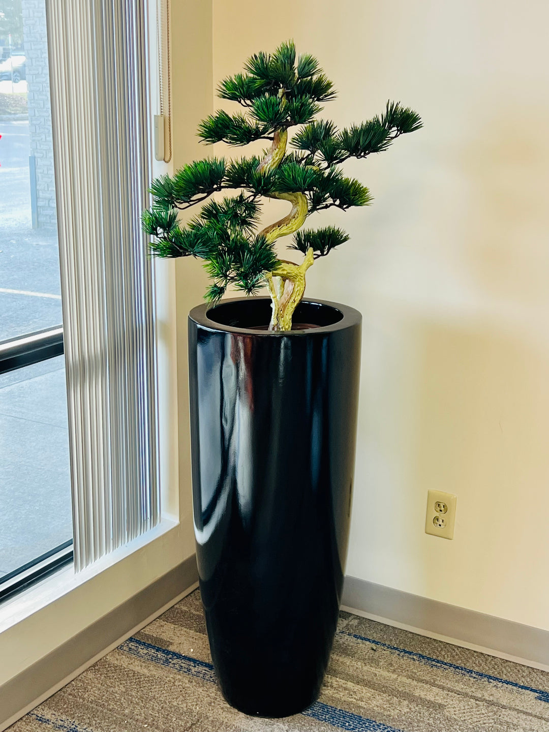 Artificial bonsai tree with a twisted trunk and lush green foliage, placed in a tall, glossy black pot, positioned near a window with vertical blinds.