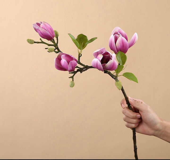 A single artificial pink magnolia branch held by a hand. The branch features vibrant pink magnolia blooms and green leaves, perfect for creating a minimalistic floral arrangement.