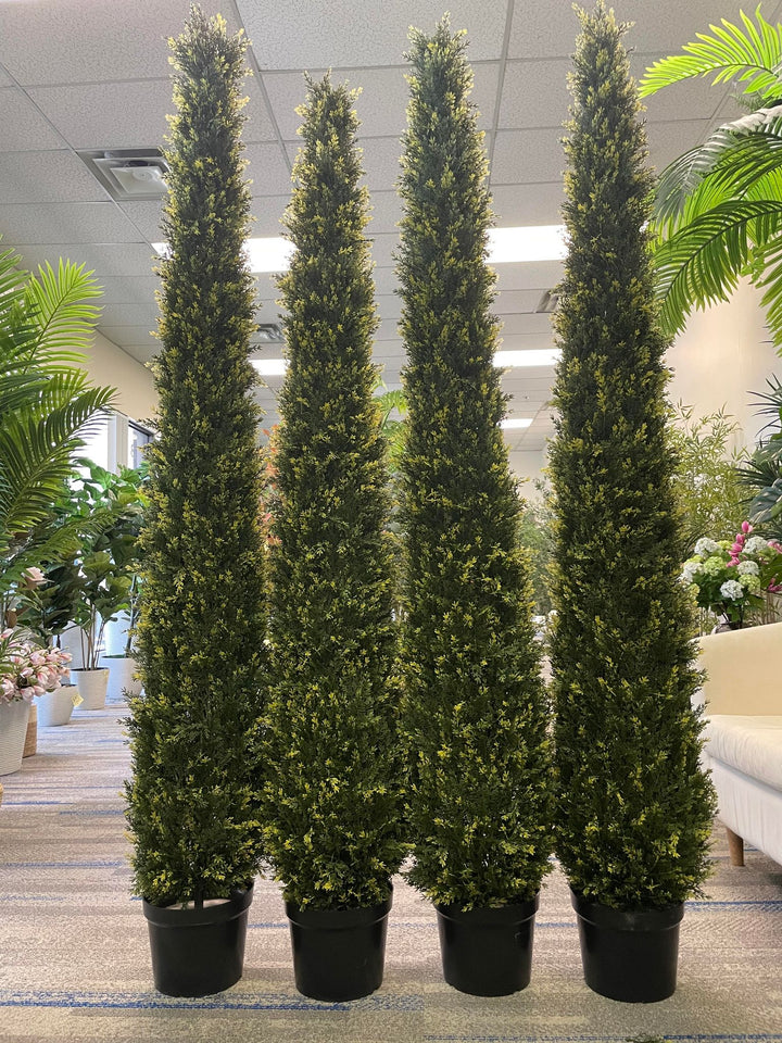 A group of tall, artificial topiaries in the shape of cones are lined up in front of a large window. The topiaries vary in height, creating a staggered arrangement. Each topiary is potted in a black planter, and the dense, leafy texture of the greenery contrasts with the smooth glass surface of the window behind them.
