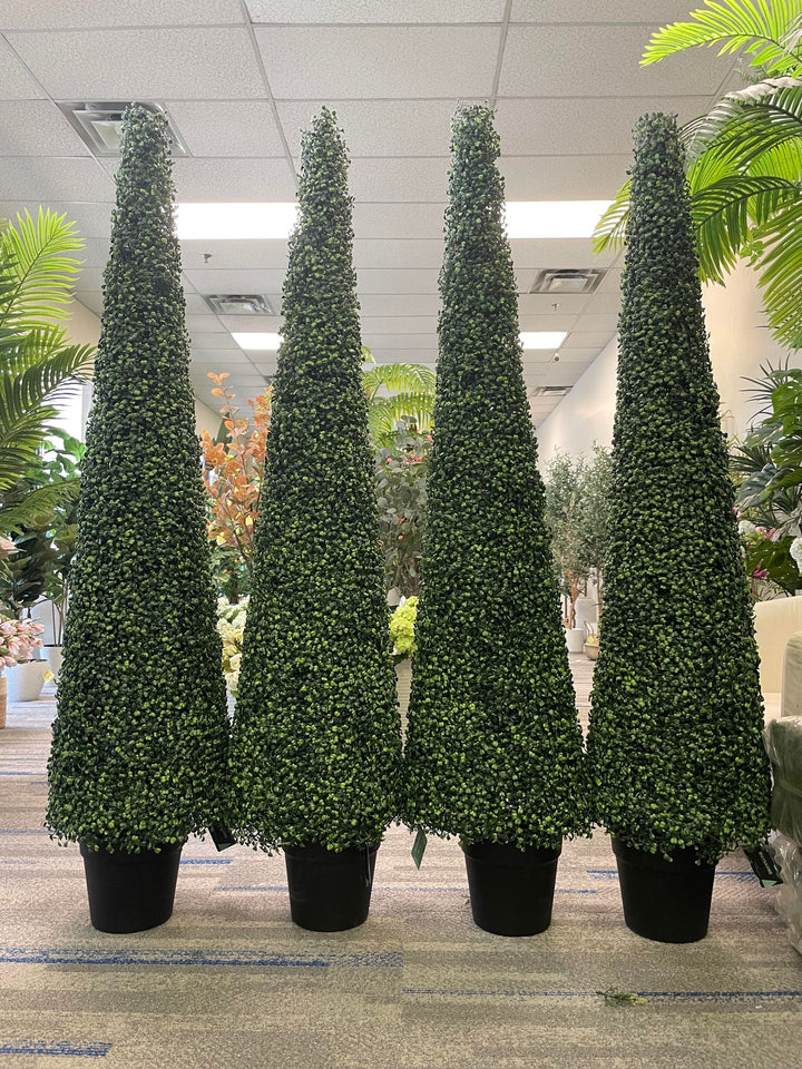Four tall, cone-shaped artificial topiary trees are arranged in a row, each in a black pot, displayed indoors with other plants in the background