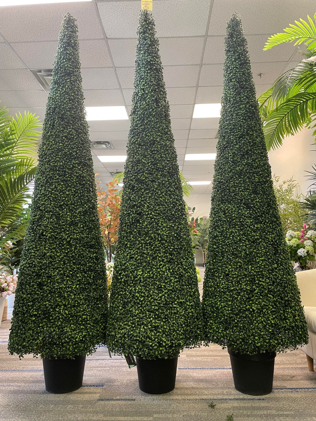 Three tall, cone-shaped artificial topiary trees are displayed in black pots within an indoor setting, surrounded by other decorative plants. The uniform height and dense foliage of the trees create a visually appealing and symmetrical arrangement.