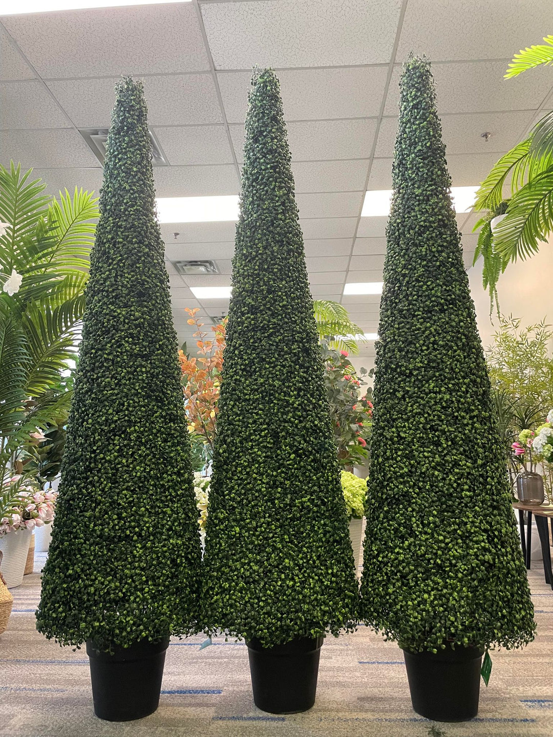 Three tall, cone-shaped artificial topiary trees are displayed in black pots within an indoor setting, surrounded by other decorative plants. The uniform height and dense foliage of the trees create a visually appealing and symmetrical arrangement