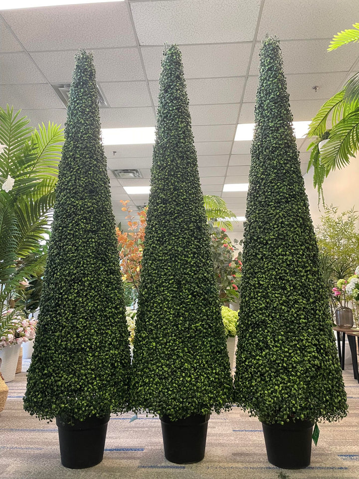 Three tall, cone-shaped artificial topiary trees are displayed in black pots within an indoor setting, surrounded by other decorative plants. The uniform height and dense foliage of the trees create a visually appealing and symmetrical arrangement